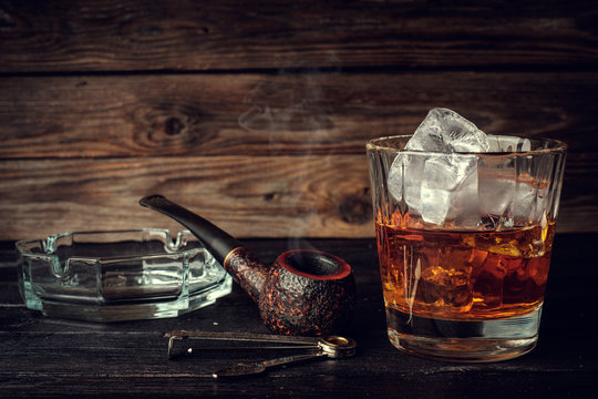 Glass of whiskey with ice and pipe on a wooden background.