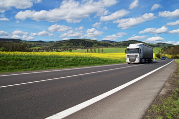White truck and private cars driving on road between flowering rapeseed field