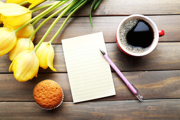 Cup of coffee with fresh cupcake, tulips and blank sheet of paper on wooden background