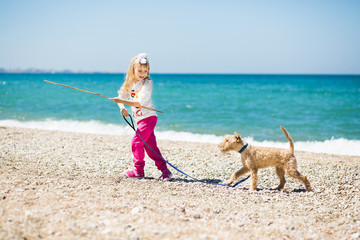 Pretty little girl with blond hair walking on the beach with a puppy terrier