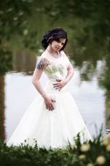 Fototapeta na wymiar Beautiful young bride in white dress near lake in summer green park
