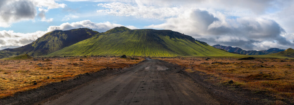 Panoramic view of dirt road leading to green, moss covered volcano mountain under a blue sky with white clouds, Landmannalaugar, Iceland.