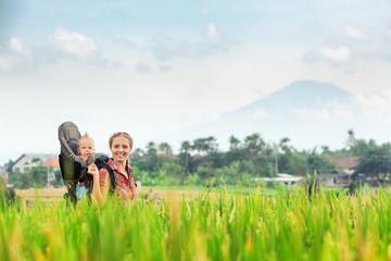 Smiling mother with baby boy in carrying backpack walking on green rice terraces. Traveling with child, active and healthy lifestyle during family summer vacation with son on Asian tropical island
