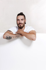 Junger Mann mit Bart und Tattoo lehnt über weißer Wand