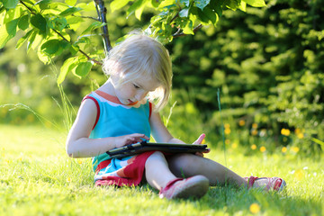 Happy preschooler child playing with tablet pc sitting in the garden on the grass. Cute little girl...