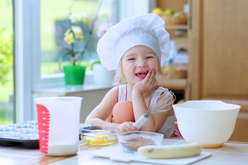 Baking with children: little happy kid, adorable toddler girl in white chef hat helping mother to...
