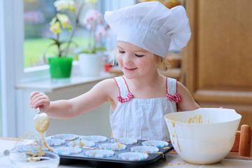 Baking with children: little happy kid, adorable toddler girl in white chef hat filling cupcakes...