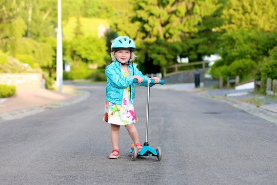 Preschooler girl riding scooter outdoors. Happy cute little child wearing blue safety helmet playing on the street learning to balance on kick board in the countryside