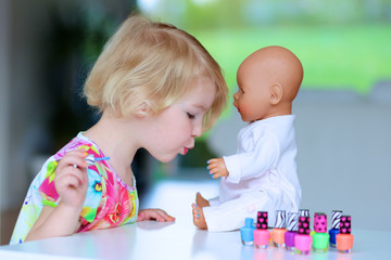 Little child, cute toddler girl having fun playing at home with colorful nail polish doing manicure and painting nails to her doll