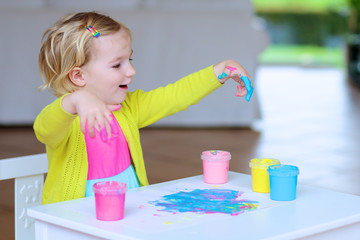 Funny little child, blonde artistic toddler girl painting with colorful finger paints indoors at bright room at home or kindergarten
