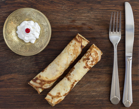 Traditional sweet pancakes on wooden plate.
Two fried folded pancakes with cheese stylish saucer decorated white cream with red berry folk and knife rough country wood background