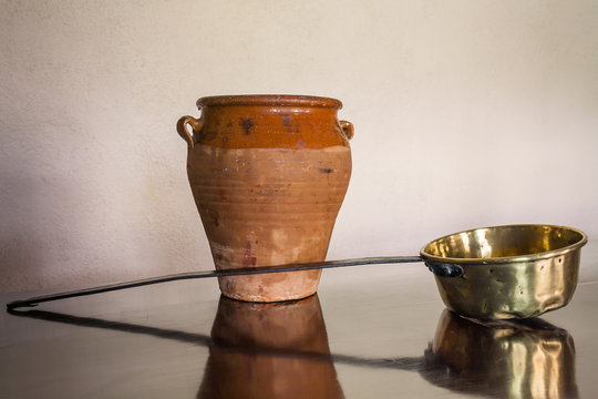 Tinaja de barro y cazo de cobre contra una pared blanca, sobre mesa brillante que los refleja