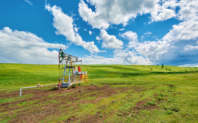 Fototapeta na wymiar Pumping unit for pumping oil on a green meadow against a blue sky with clouds in summer
