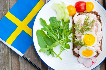 Swedish style rye bread open sandwich topped with prawns and boiled egg. Served on a rustic wooden table with a side salad of rocket leaves, tomato and cucumber and garnished with fresh dill.
