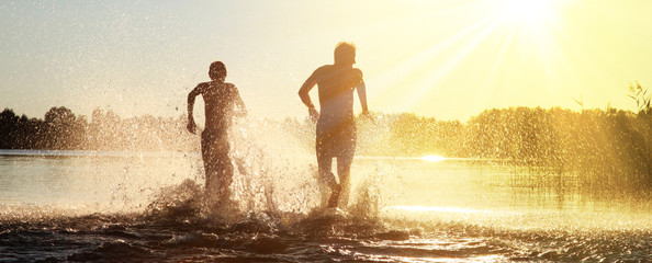 Glückliche junge Menschen laufen und springen am See beim Sonnenuntergang - obrazy, fototapety, plakaty