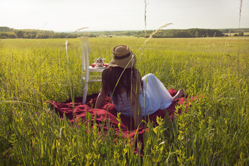 Woman with Hat in White Dress is Sitting on Red Cloth on Green Meadow with WHite Chair and Picnic...