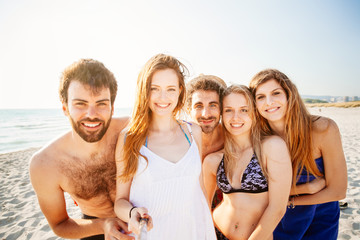 Fototapeta premium Selfie on the beach! Five friends in swimwear (two men and three women) take a photo at the sea in a summer day at sunset on the beach with no other people