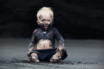 Funny portrait of smiling child with dirty face sitting and playing with fun on black sand sea...