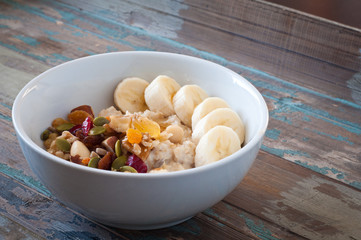 Bowl of freshly prepared home made porridge oatmeal topped with sliced banana and dried fruit, nuts and seeds. A healthy breakfast served on a rustic wooden tray.