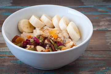 Fototapeta premium Bowl of freshly prepared home made porridge oatmeal topped with sliced banana and dried fruit, nuts and seeds. A healthy breakfast served on a rustic wooden tray.