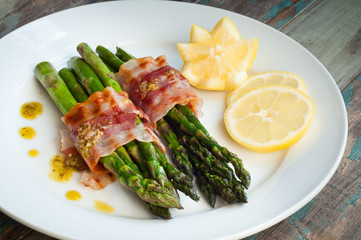 Fresh grilled asparagus wrapped in pancetta bacon served with a drizzle of pesto and freshly cut lemon. Served on a rustic wooden table.
