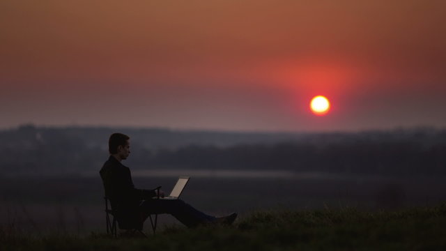5 in 1 video! The man sit and work on the laptop by sunset (sunrise) background on the top of the hill. VIDEO WITH VARIABLE PLAYBACK SPEED