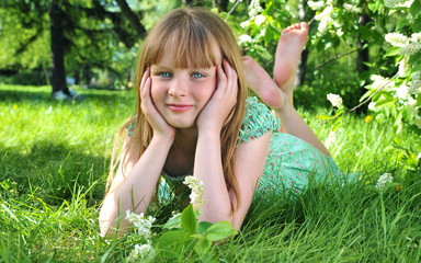 Naklejka na ściany i meble Little girl lies on green grass on background white flowers of bird cherry 