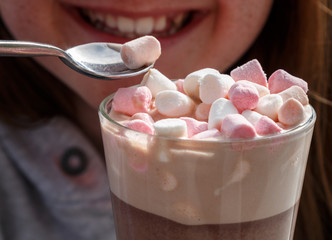 Marshmallows on the top of a hot chocolate drink in a glass being eaten by a young smiling pretty...
