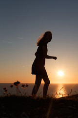 Teenage girl walking along cliff edge looking out to sea in silhouette from the sun setting into the sea.