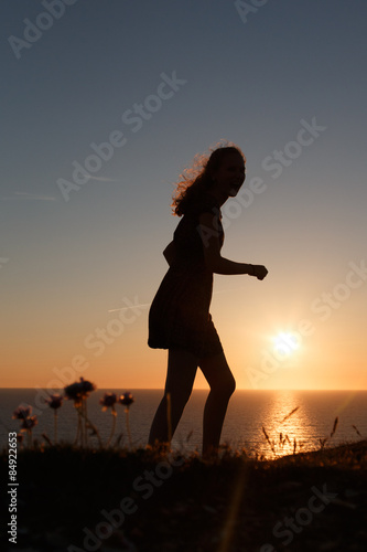 Teenage Girl Walking Along Cliff Edge Looking Out To Sea In Silhouette From The Sun Setting Teenage Girl Walking Along Cliff Edge Looking Out To Sea In Silhouette From The Sun Setting