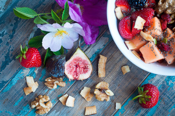 Bowl of home made fresh fruit muesli topped with strawberries,melon,blackberries,walnuts,chia seeds,peanut butter and walnuts. Served on a rustic wooden table with fresh wild flowers.
