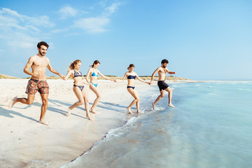 A summer day on a sandy beach, a group of five young people run towards the sea to swim. People are in swimwear on the beach and there are no other people.