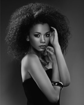 Beautiful young African American woman with afro in a fresh dark short summer dress posing holding up one edge of the flared skirt with a provocative expression, on a dark grey background