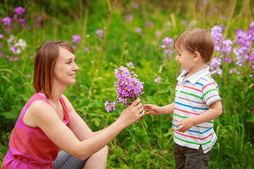 Portrait of mother and son together sitting in green grass on meadow field  in countryside smiling holding smelling flowers, happy family concept, summer fun activity, Mothers Day