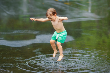 Portrait of happy toddler boy jumping in puddles during the rain thunderstorm on a bright summer day outside, sports recreation leisure concept, childhood and freedom