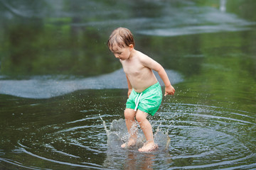 Portrait of happy toddler boy jumping in puddles during the rain thunderstorm on a bright summer day outside, sports recreation leisure concept, childhood and freedom