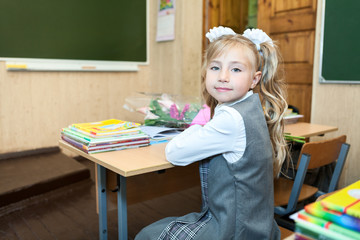 Cute schoolgirl in school uniform turning back while sitting at desk in classroom