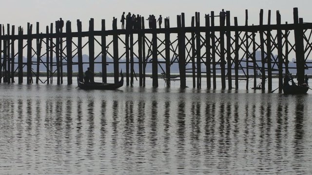 Silhouettes of people and pillars of U Bein bridge reflect in water of lake