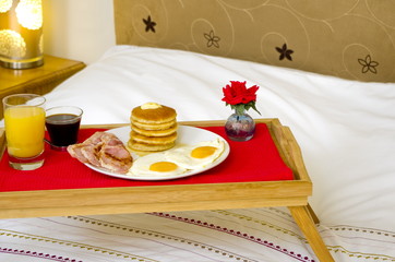 A  tray with a Pancake Maple Syrup, bacon and egg breakfast on a double bed.