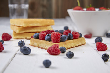 Different berries on a waffle and in a bowl on a white wooden table