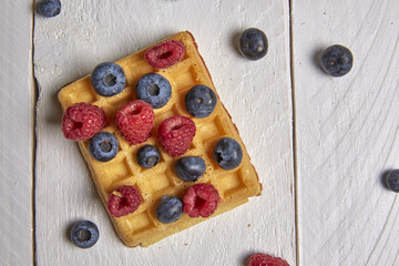 Different berries on a waffle and in a bowl on a white wooden table