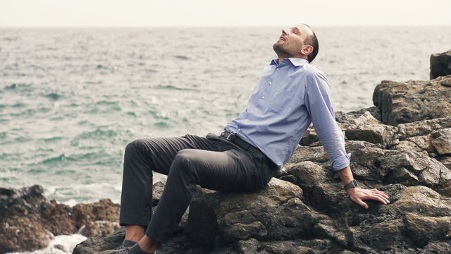 Happy businessman relaxing while sitting on rocks near sea, slow motion shot at 240fps
