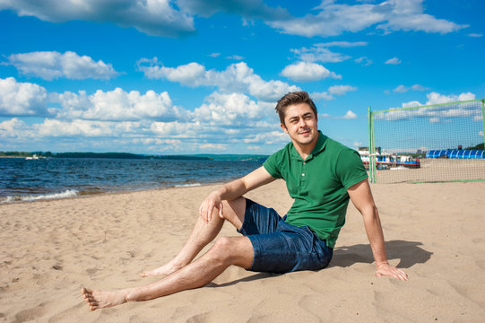 Young fit man wearing green polo sits on a sand at the beach on sunny day under scenic blue cloudy sky