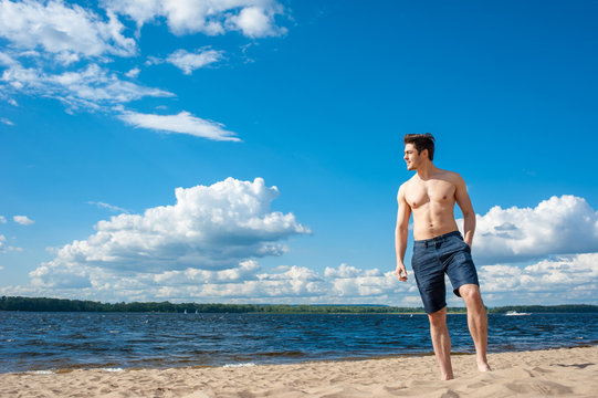 Fit young man with bare torso standing at the beach under scenic blue sky on sunny day