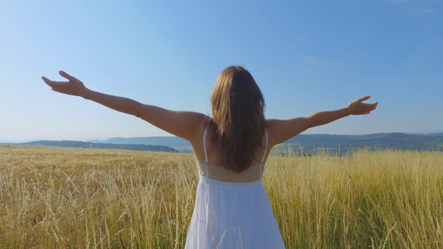 Beautiful young woman with a long white dress standing in a field of long grass lifting her arms up with joy of being surrounded  by natural beauty.