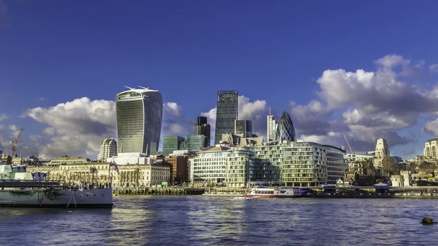 Time lapse of the City of London skyline before sunset. Dramatic cloud on a blue sky, reflections on office buildings and on the river Thames and boats passing through