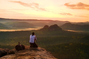 Fototapeta premium Professional photographer in white jacket takes photos with camera on tripod on rocky peak. Dreamy fogy landscape, spring orange pink misty sunrise in a beautiful valley below