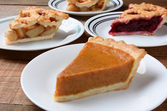Closeup of four slices of pie on dessert plates. Focus is on the front slice of pumpkin pie. The back plates have apple and cherry pie. 