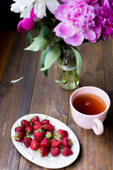 white and pink peonies, black tea and fresh strawberries from farmers market on a dark wood table in rustic style