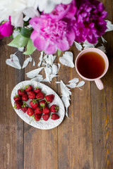 white and pink peonies, black tea and fresh strawberries from farmers market on a dark wood table in rustic style